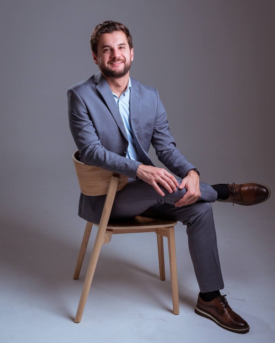Dr. Paulo Othávio Almeida sentado em uma cadeira de madeira, de terno cinza e camisa azul-clara, sorrindo